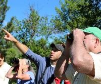 A hike leader points to a bird while a man with binoculars looks in that direction
